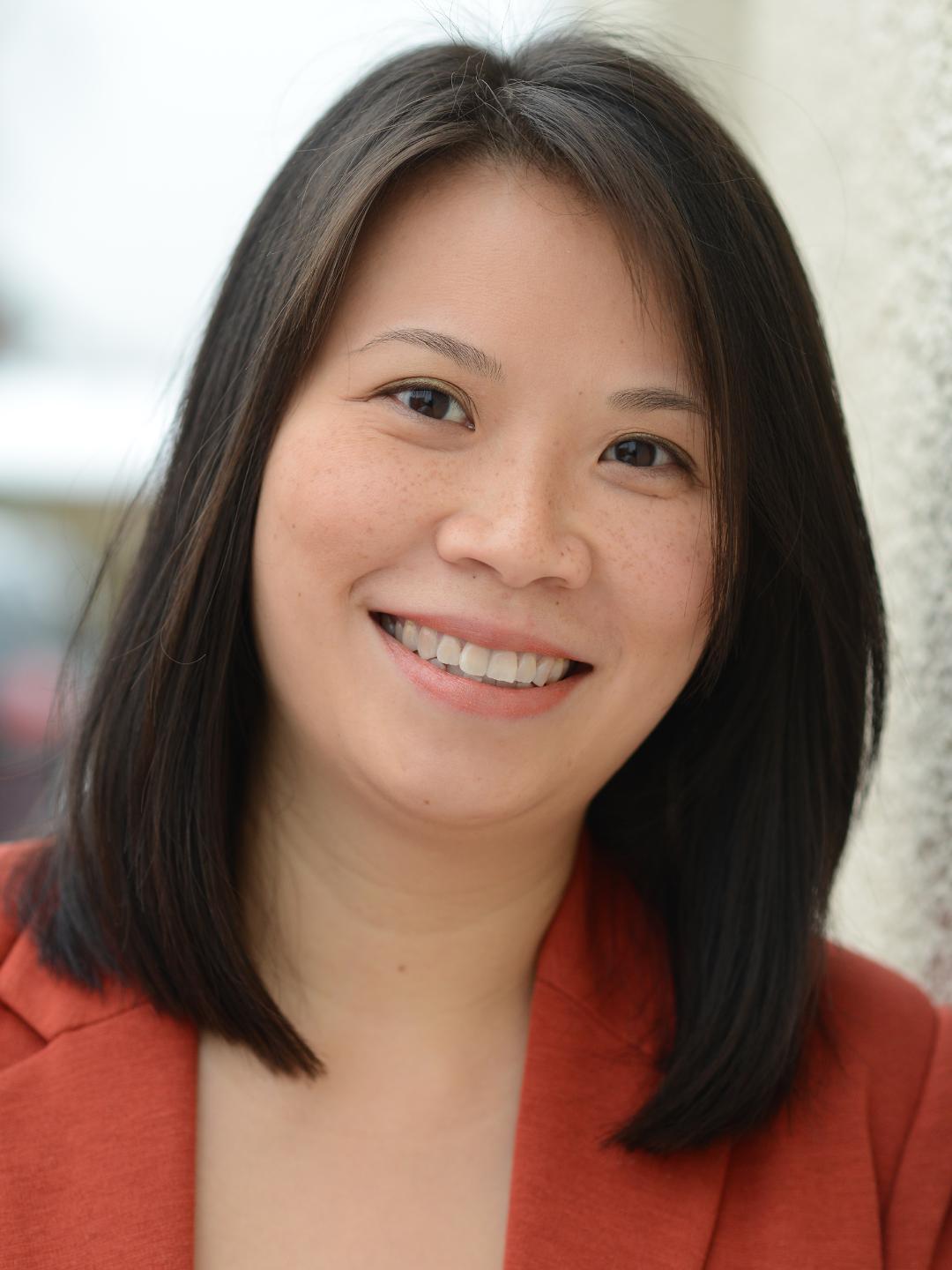 A head shot of Sharon. She has shoulder length dark brown hair. She is smiling and wearing a red jacket. 